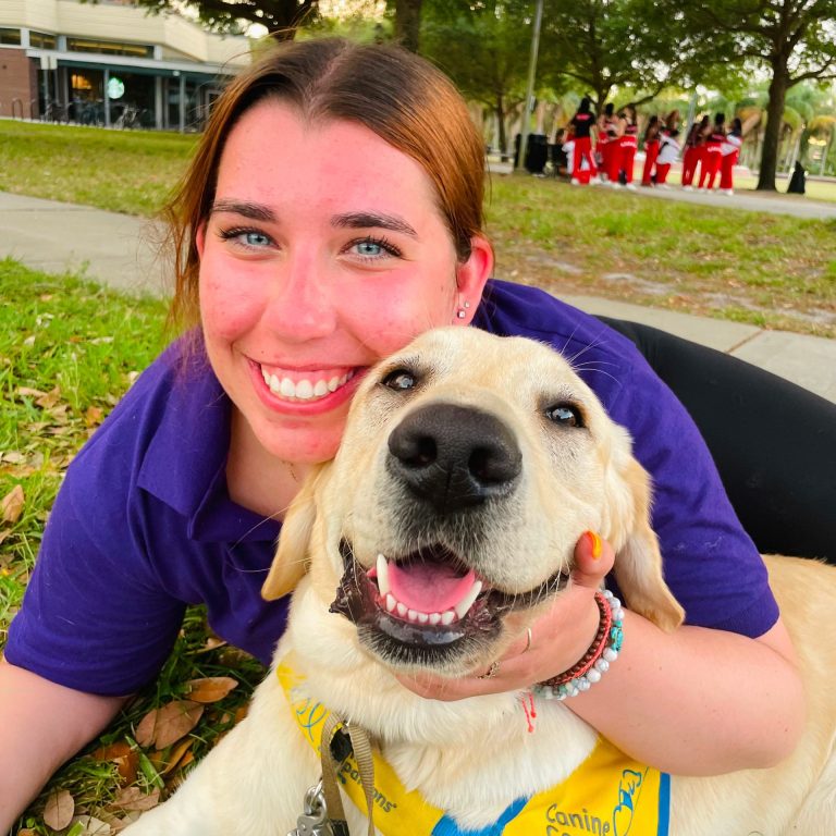 A woman in a purple shirt smiles while holding a yellow Labrador retriever wearing a “Canine Companions” bandana, both lying on grass in a park.