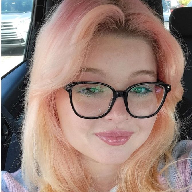 Person with light pink hair, wearing black-framed glasses and light makeup, is sitting in a car and smiling slightly at the camera.