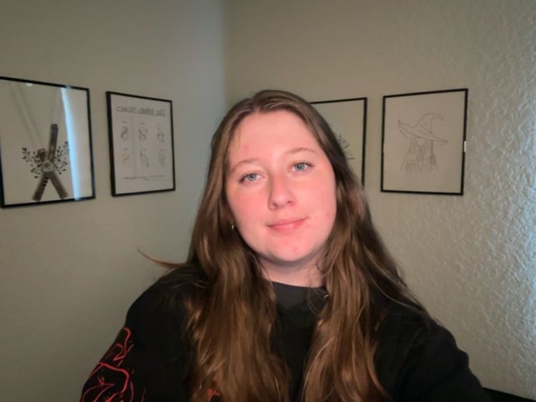 A young woman with long brown hair is looking at the camera and smiling slightly. She is indoors with framed artwork on the wall behind her.