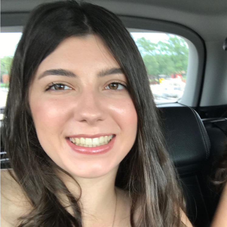A young woman with long brown hair smiles while sitting in the back seat of a car. Trees and street signs are visible through the window behind her.