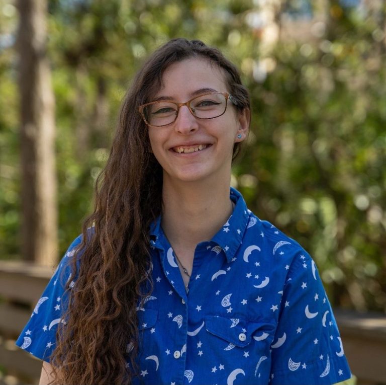 A person with long curly hair and glasses smiles outdoors, wearing a blue shirt with moon and star patterns.