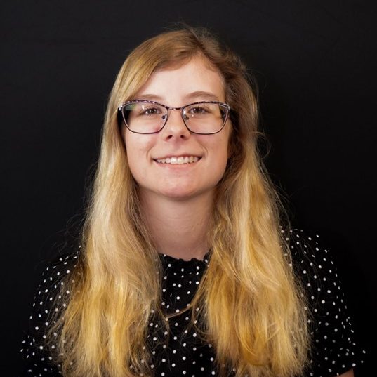 A young woman with long blonde hair, wearing glasses and a black polka dot shirt, stands in front of a plain black background, smiling at the camera.