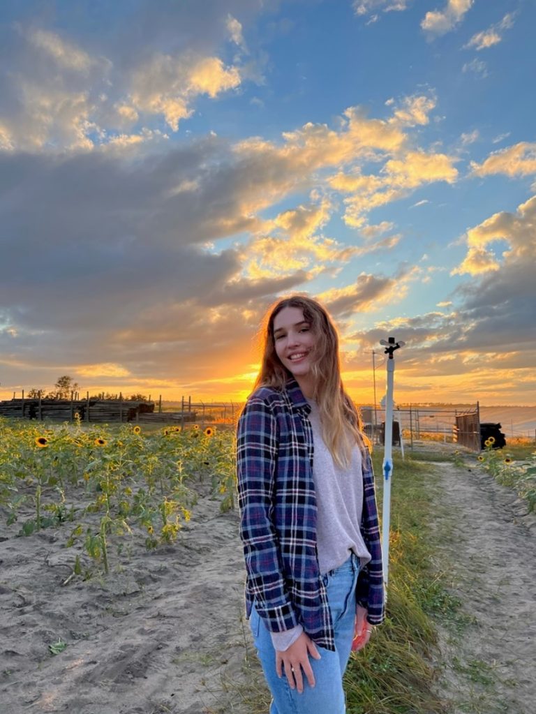 A person with long hair and a plaid shirt stands in a sunflower field at sunset, smiling at the camera. The sky is partly cloudy with warm sunlight in the background.