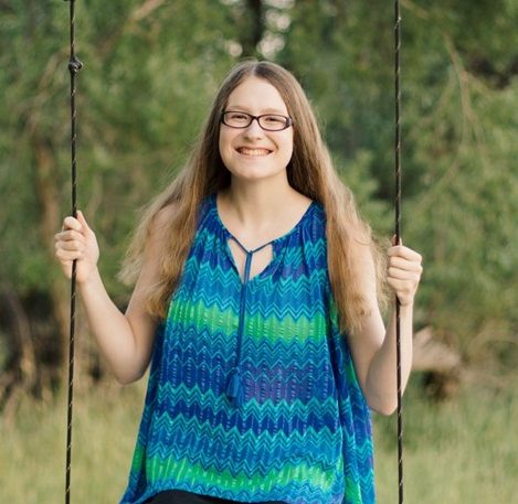 A person with long hair and glasses sits on a swing outdoors, smiling at the camera; green trees are visible in the background.