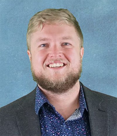 A man with short blond hair and a beard is smiling, wearing a dark blazer over a patterned shirt, posed in front of a blue background.
