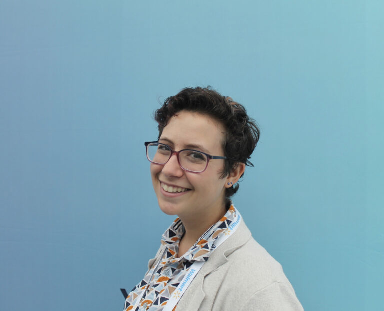 A person with short curly hair and glasses smiles at the camera, wearing a patterned shirt, light blazer, and a lanyard with "Walmart" text, in front of a plain blue background.
