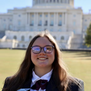 A young woman with long brown hair, glasses, and a suit stands smiling in front of a government building.