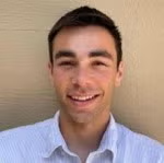 A young man with short brown hair smiles at the camera, wearing a white collared shirt and standing in front of a beige wall.