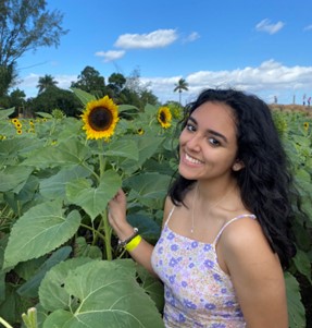 A young woman with long dark hair stands among tall sunflowers, smiling at the camera under a blue sky with scattered clouds.