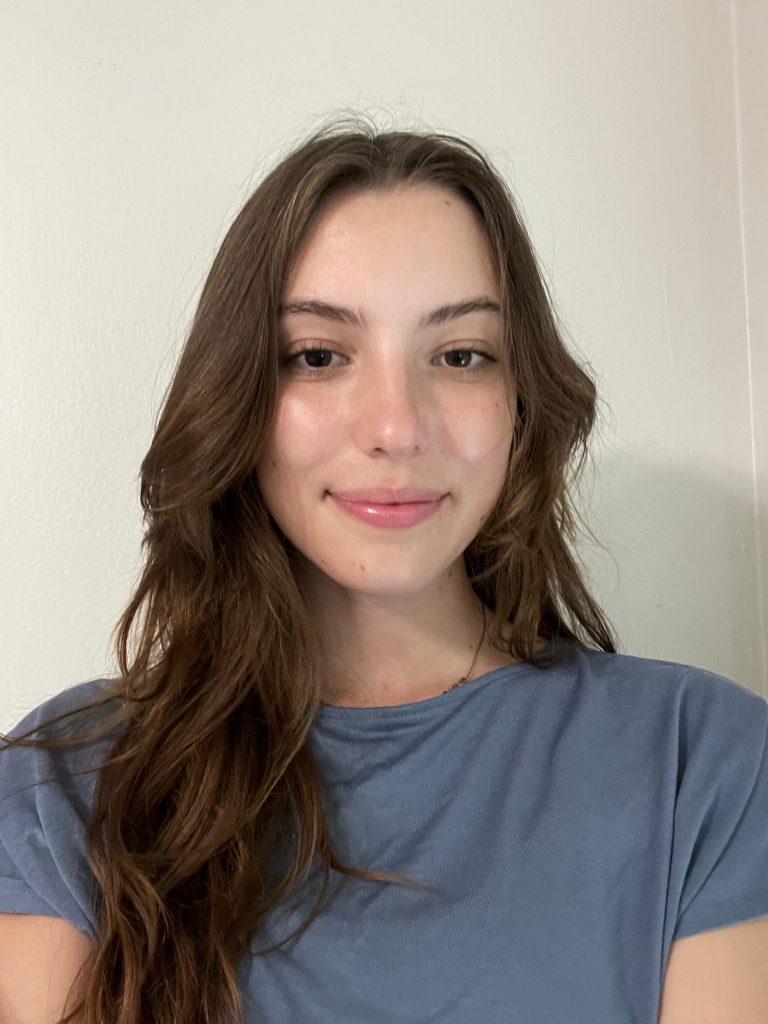 A young woman with long brown hair and a blue shirt stands in front of a plain light-colored wall, looking at the camera with a neutral expression.