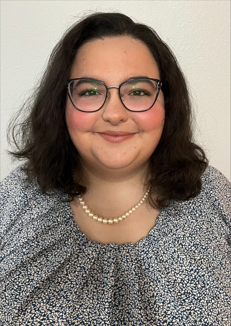 A woman with shoulder-length dark hair, glasses, and a pearl necklace smiles at the camera. She is wearing a patterned blouse and is posed against a plain light-colored wall.