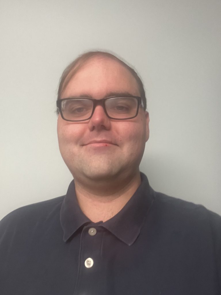 A man wearing glasses and a dark polo shirt stands against a plain light-colored wall, looking at the camera with a neutral expression.