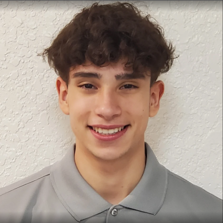 A young person with curly brown hair, wearing a light gray collared shirt, smiles in front of a white textured wall.
