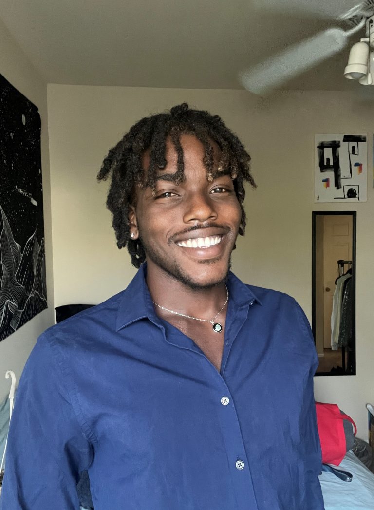 A person with short dreadlocks, wearing a blue button-up shirt and a necklace, smiles while standing in a bedroom with artwork and a ceiling fan visible.