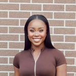A woman with straight, long black hair and a brown zip-up top stands smiling in front of a brick wall.