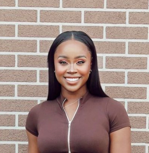 A woman with straight, long black hair and a brown zip-up top stands smiling in front of a brick wall.