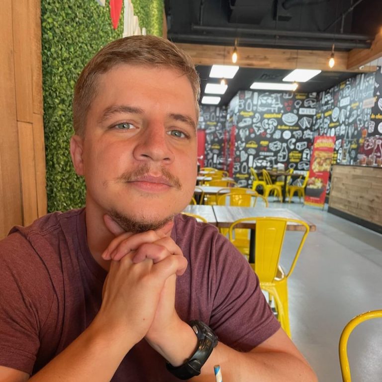 A man sits at a table in a casual restaurant with yellow chairs, arms crossed in front of his face, facing the camera. The background features doodle art on the walls.