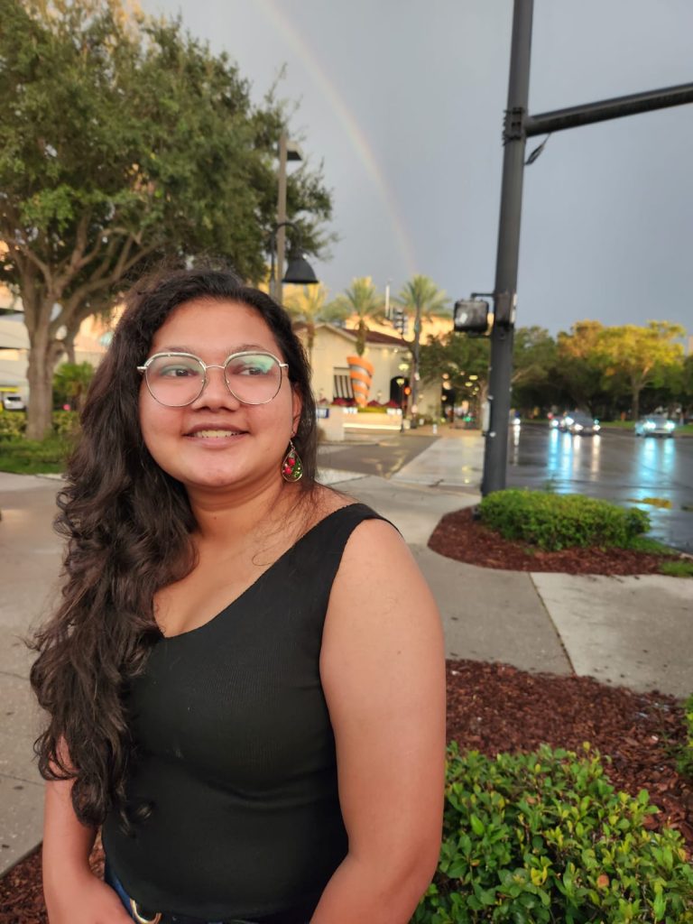 A woman with long hair and glasses stands on a city sidewalk at dusk, with a rainbow visible in the sky behind her.