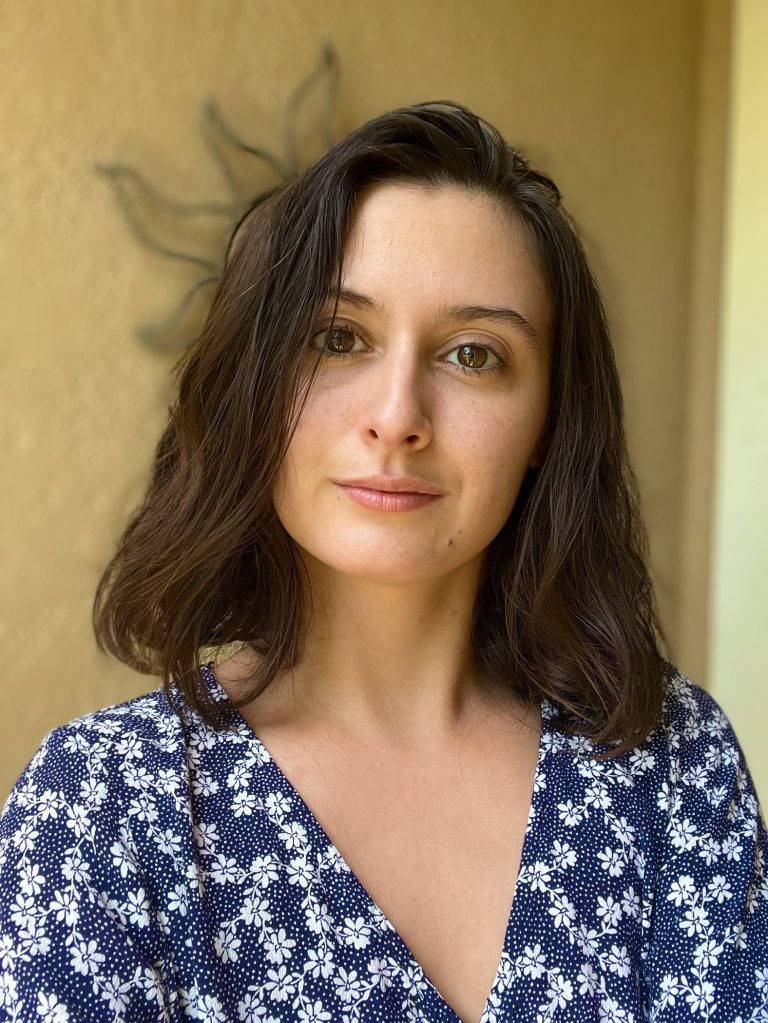 A woman with shoulder-length brown hair wearing a blue and white floral top stands in front of a beige wall with a sun-shaped decoration in the background.