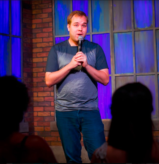 A man holding a microphone performs stand-up comedy on stage in front of an audience, with blue and purple lighting and brick walls in the background.