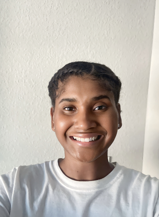 A person with short braided hair smiles at the camera, wearing a white shirt, standing in front of a light-colored textured wall.