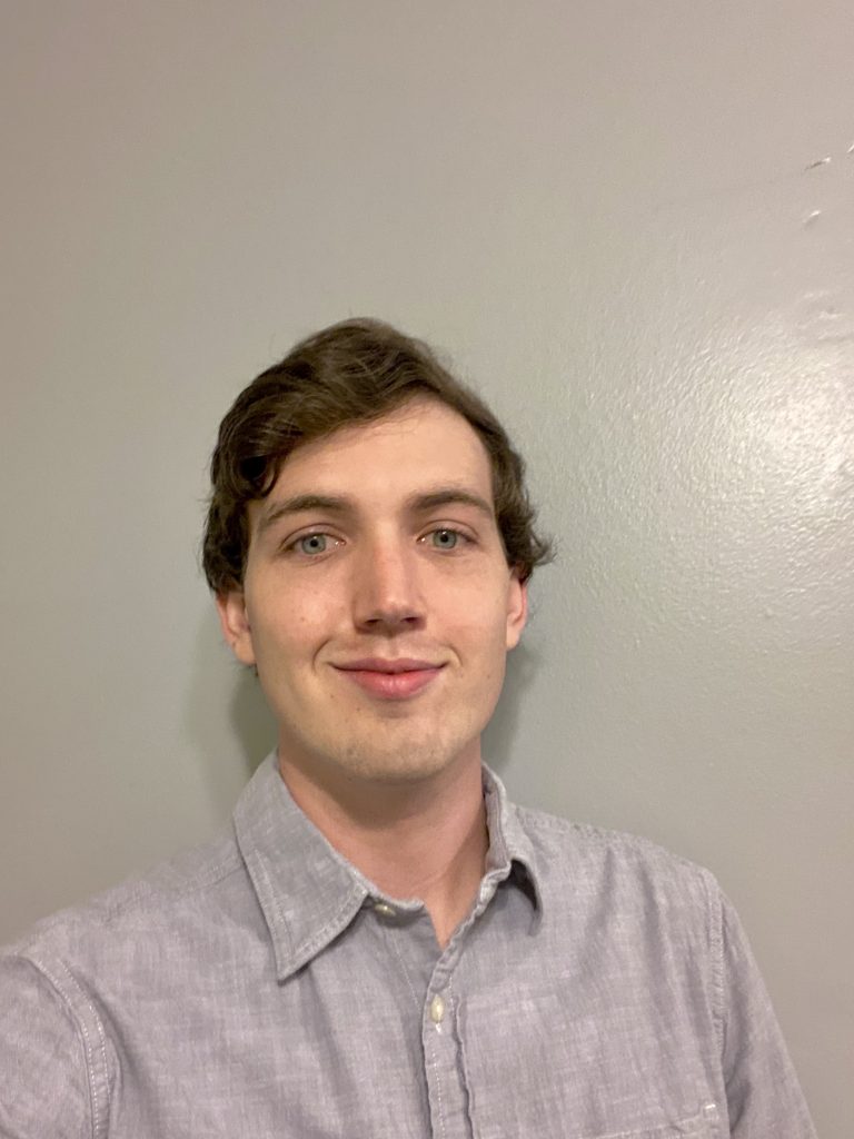 A young adult wearing a light gray collared shirt stands in front of a plain light gray wall, looking at the camera and smiling slightly.