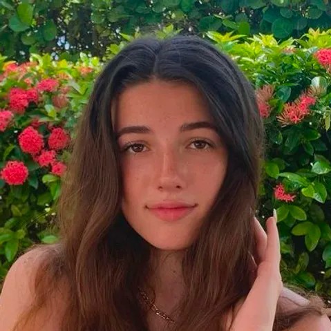 A young woman with long brown hair poses outdoors in front of green bushes with pink flowers.