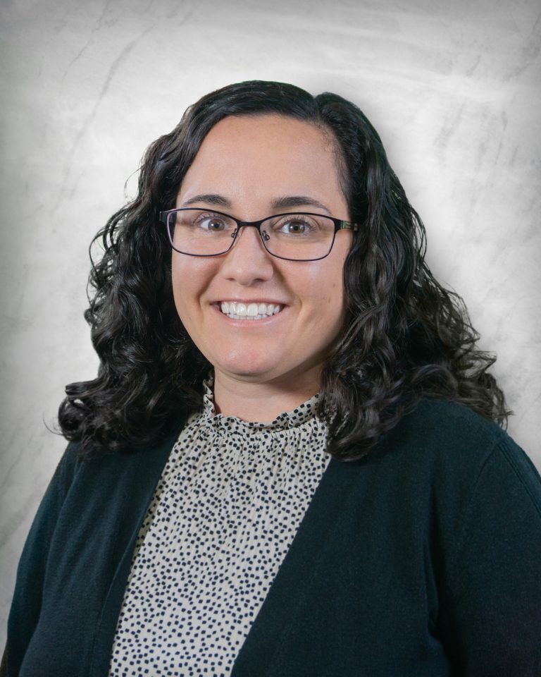A woman with curly dark hair and glasses, wearing a black cardigan over a polka-dotted blouse, smiles in front of a light gray textured background.