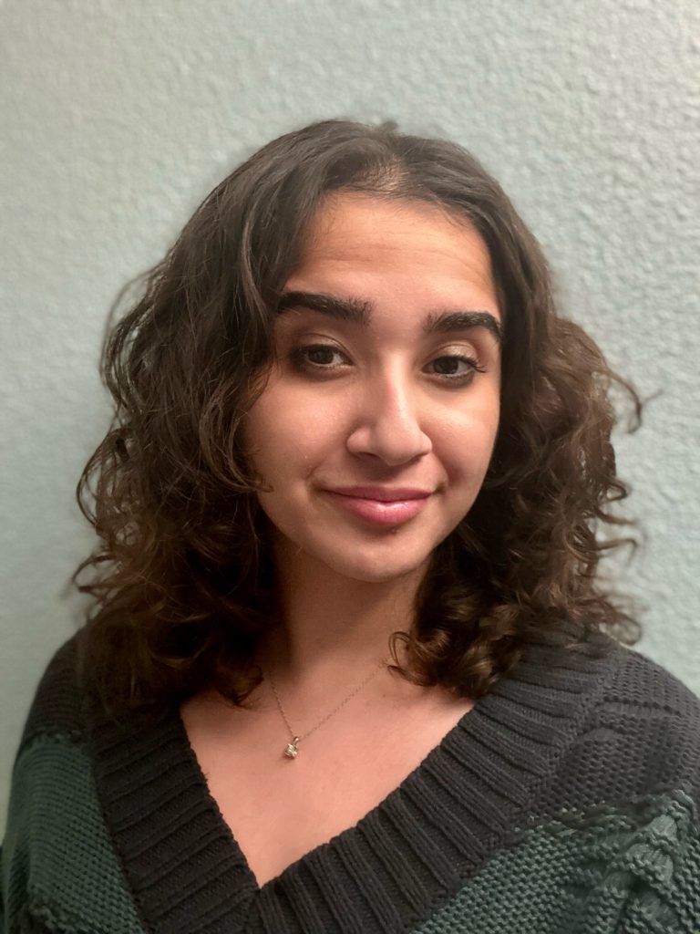 A woman with curly brown hair wears a dark sweater and a small necklace, standing in front of a light-colored textured wall.
