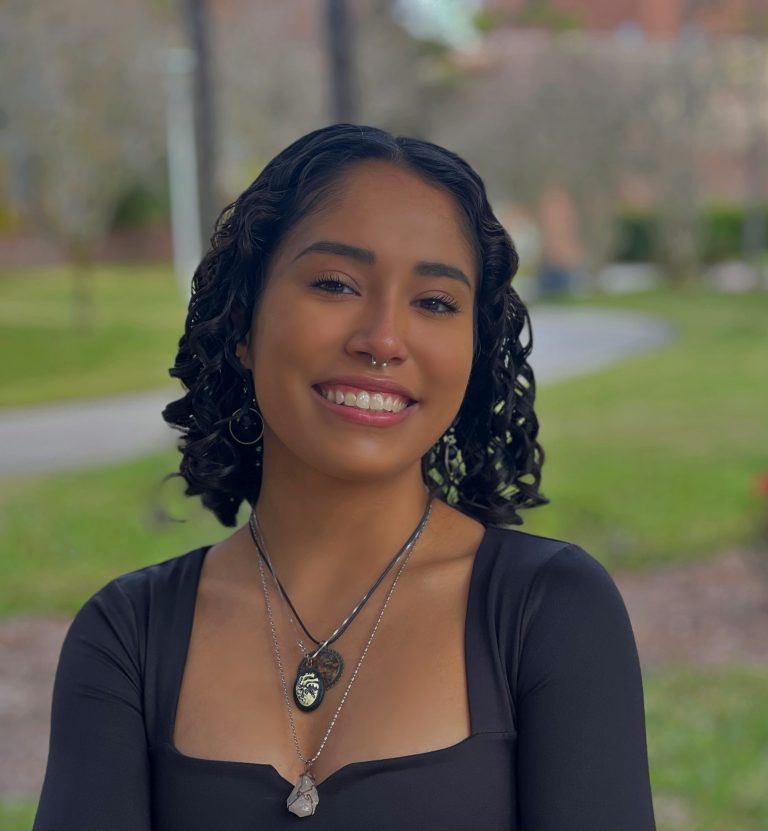 A young woman with curly hair wearing a black top and layered necklaces smiles outdoors in a park-like setting.