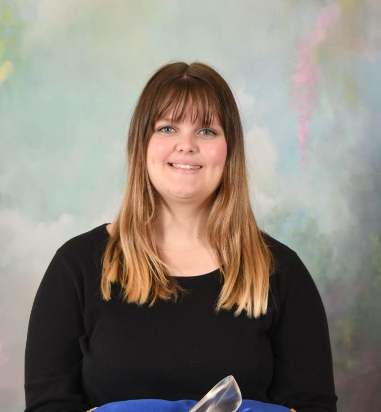 A woman with straight, shoulder-length hair and bangs, wearing a black top, sits in front of a pastel-colored backdrop, smiling at the camera.