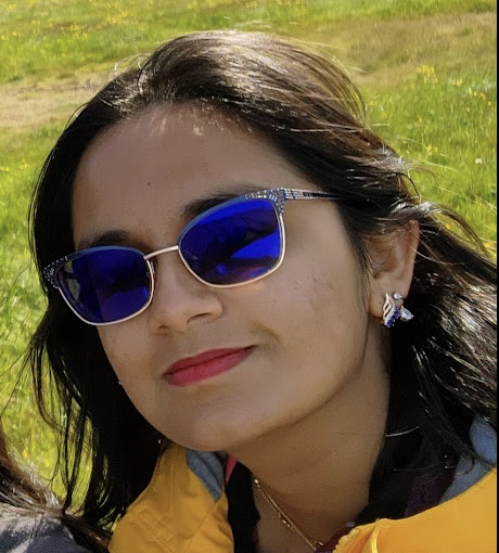 A woman wearing blue reflective sunglasses, a yellow jacket, and butterfly-shaped earrings stands outdoors in a grassy area.