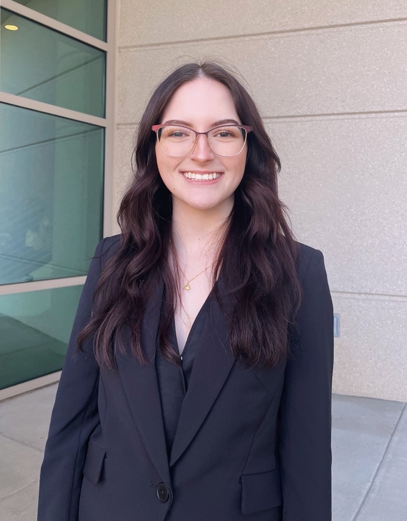 A woman with long brown hair and glasses, wearing a black blazer, stands and smiles in front of a beige building with glass windows.