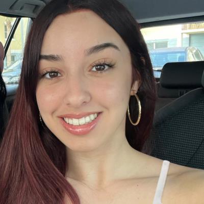 A young woman with long dark hair and hoop earrings smiles at the camera while sitting in a car.