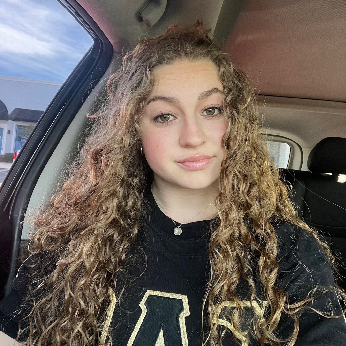 A young woman with long, curly hair sits in a car, wearing a black T-shirt with a large "A" on it, looking directly at the camera with a neutral expression.