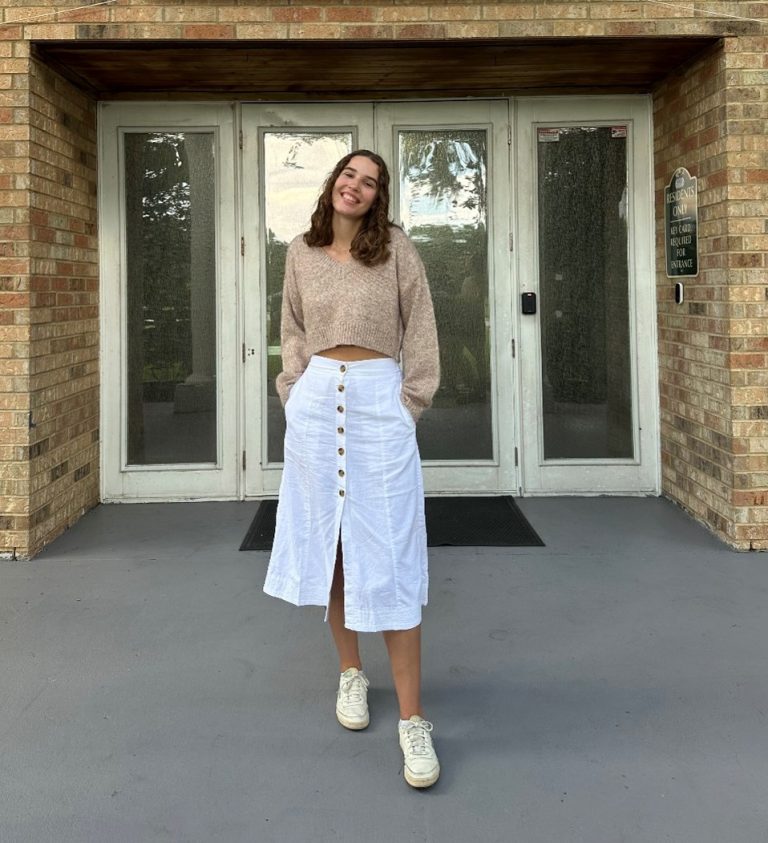 A woman stands in front of glass doors, wearing a beige sweater, white button-front skirt, and white sneakers, with her hands in her skirt pockets.