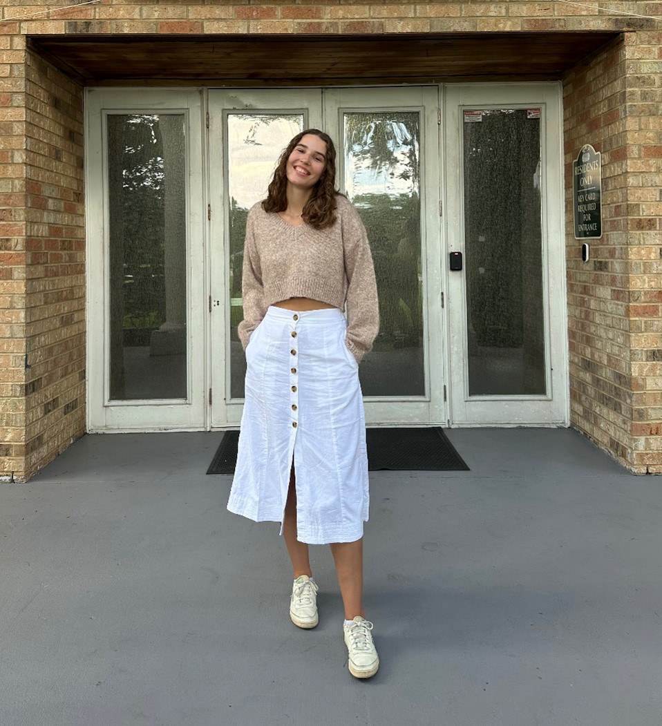 A woman stands in front of glass doors, wearing a beige sweater, white button-front skirt, and white sneakers, with her hands in her skirt pockets.