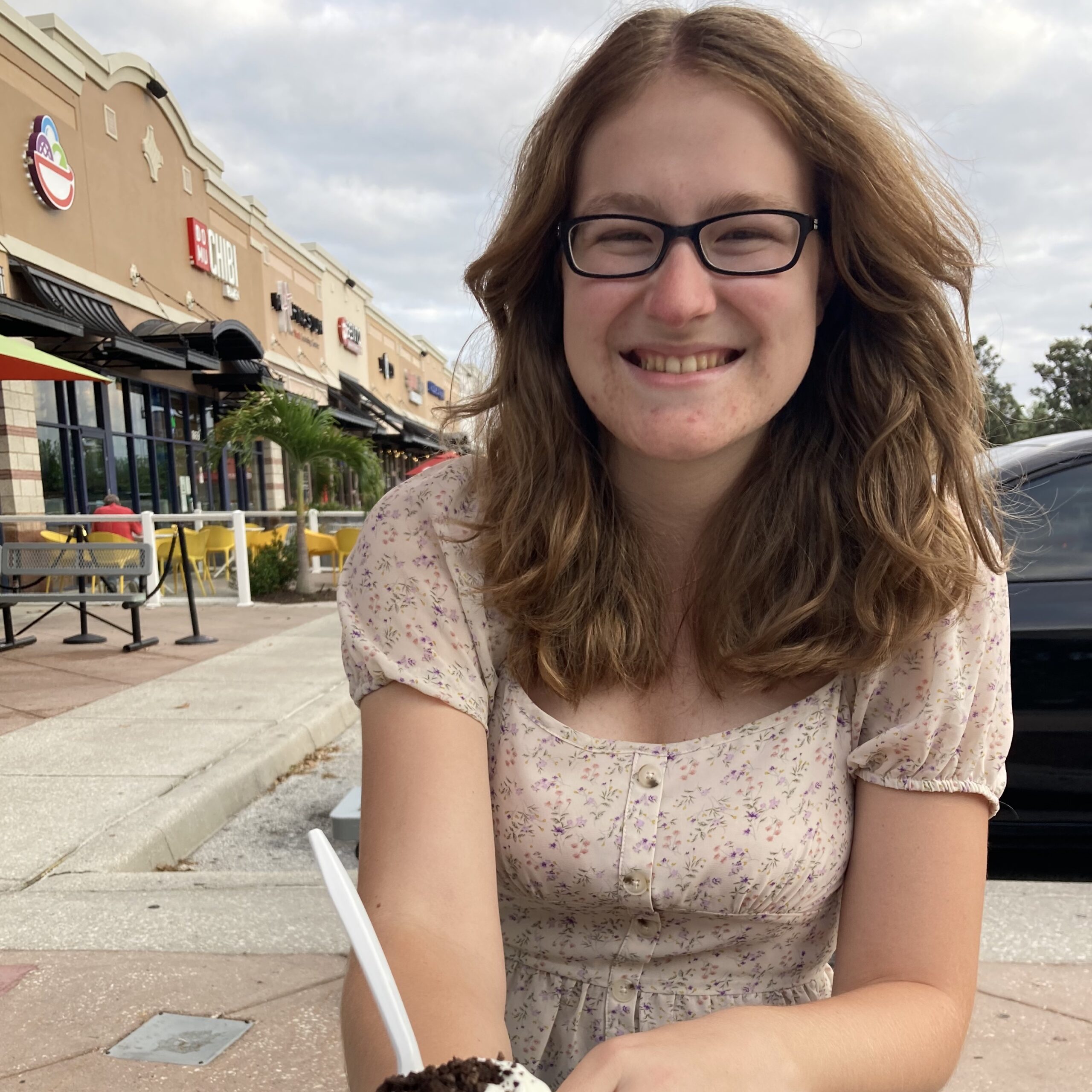A young woman with glasses and wavy brown hair smiles at an outdoor seating area, holding a cup of dessert. Shops and benches are visible in the background.