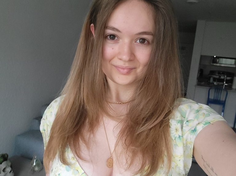 A woman with long brown hair, wearing a floral dress and two necklaces, smiles at the camera indoors.