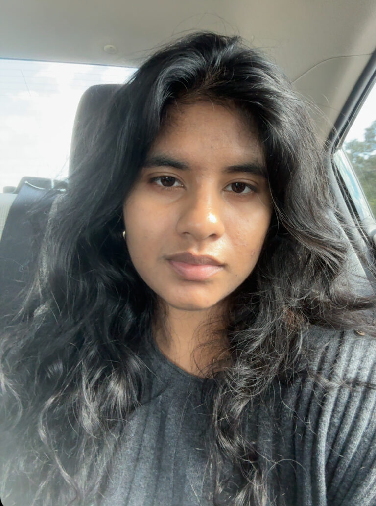 A person with long, wavy black hair and a neutral expression sits in a car, wearing a dark ribbed top. Sunlight filters through the windows.