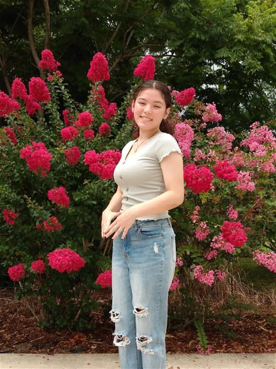 A young woman in a light green shirt and ripped jeans stands smiling in front of vibrant pink flowering bushes and green trees.