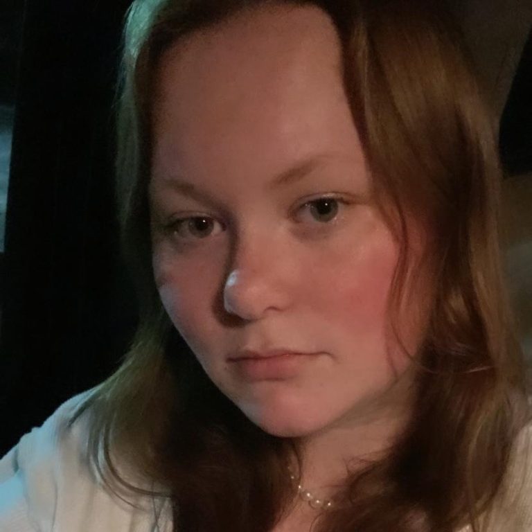 A person with fair skin and long, light brown hair looks at the camera with a neutral expression, sitting indoors with dim lighting.