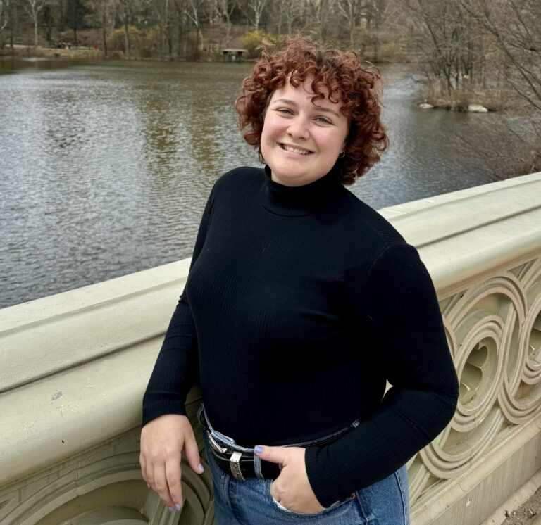 A person with curly hair wearing a black turtleneck and jeans stands smiling on a bridge overlooking a lake and trees.