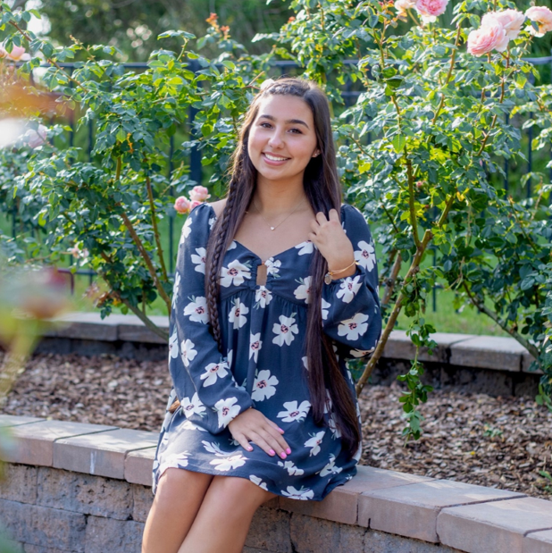 A young woman with long dark hair, wearing a blue floral dress, sits on a stone bench in a garden with blooming pink roses.