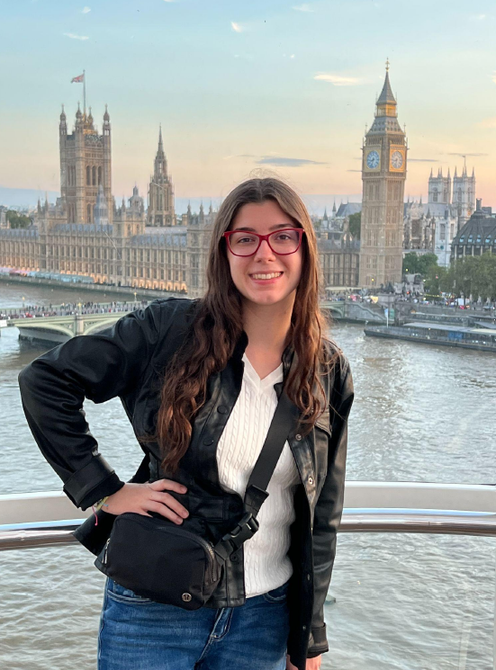 A woman wearing glasses and a black jacket stands in front of the River Thames with the Houses of Parliament and Big Ben in the background at sunset.