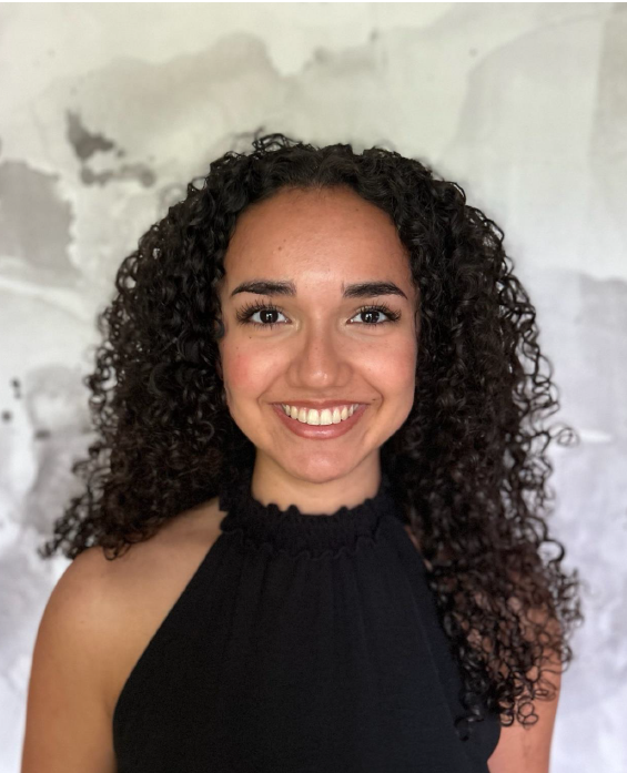 A woman with curly dark hair and a sleeveless black top smiles at the camera against a light, abstract background.