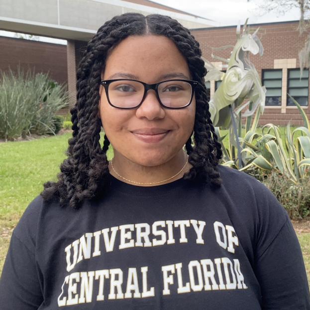 A person wearing glasses and a "University of Central Florida" t-shirt stands outside near a building and plants, facing the camera and smiling.