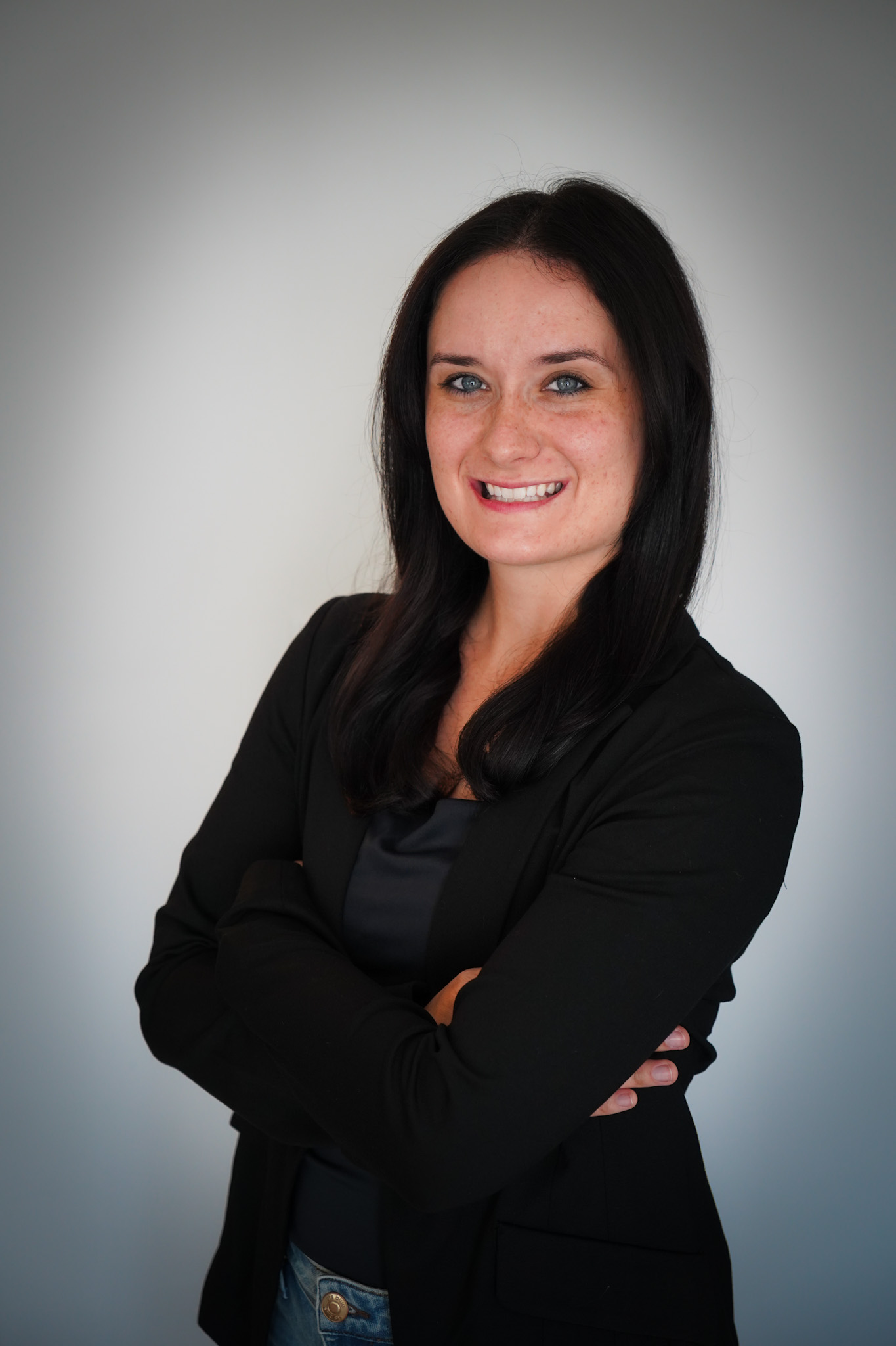 Woman with long dark hair wearing a black blazer and dark top standing against a plain background, smiling with arms crossed.