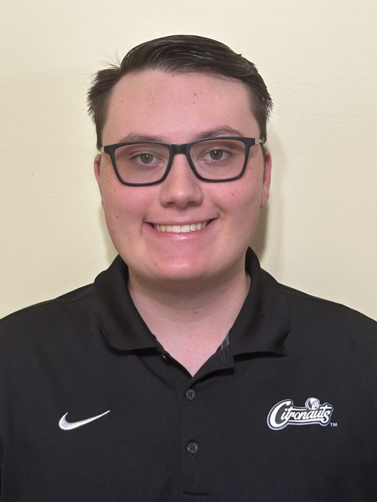 A person with short dark hair and glasses smiles at the camera, wearing a black Nike Cincinnati polo shirt against a light-colored background.