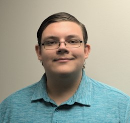 A person wearing glasses and a teal collared shirt is facing the camera and smiling slightly, with a plain light-colored background.